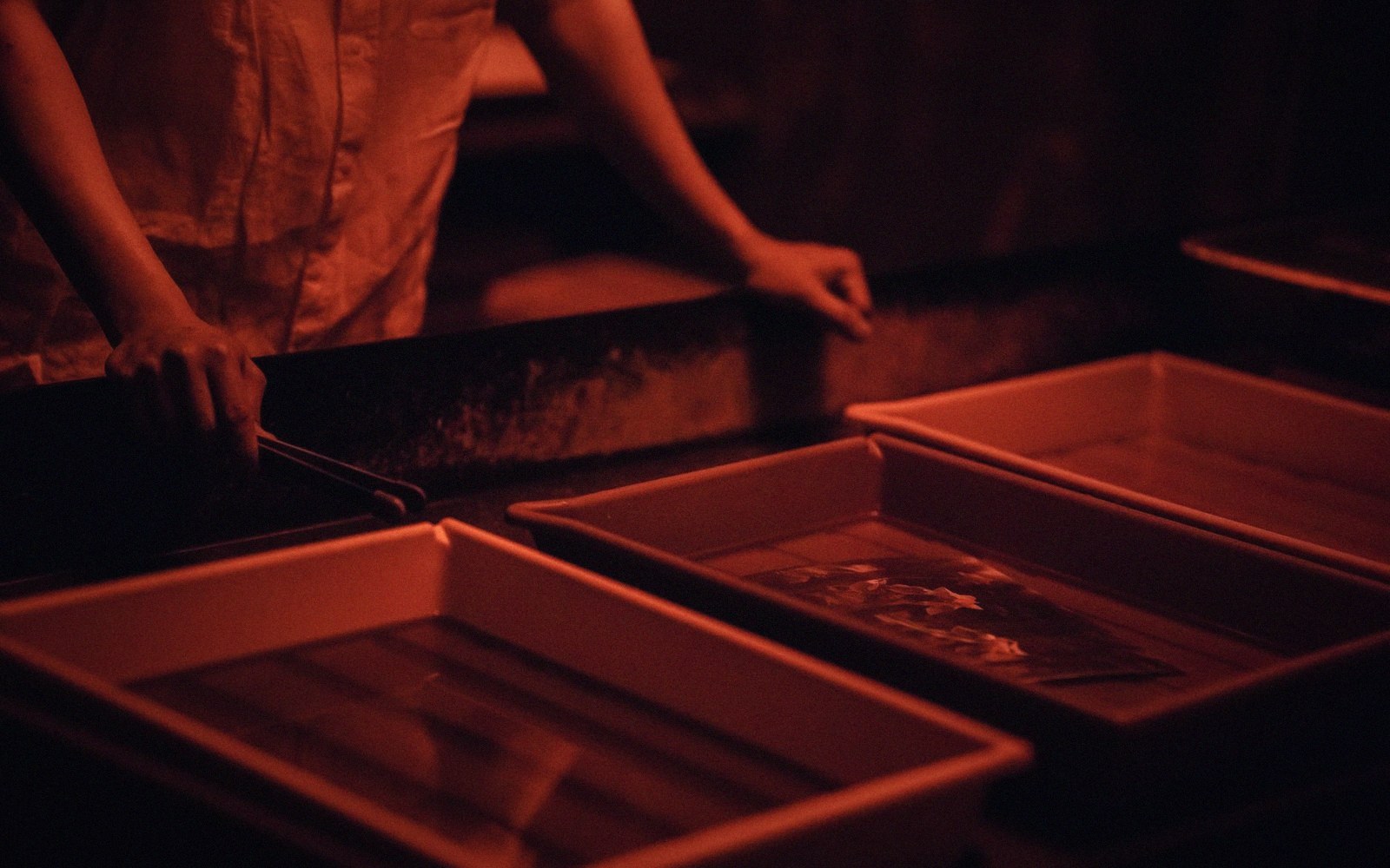 A person leans over trays of developing photographs in a darkroom lit by red safelight