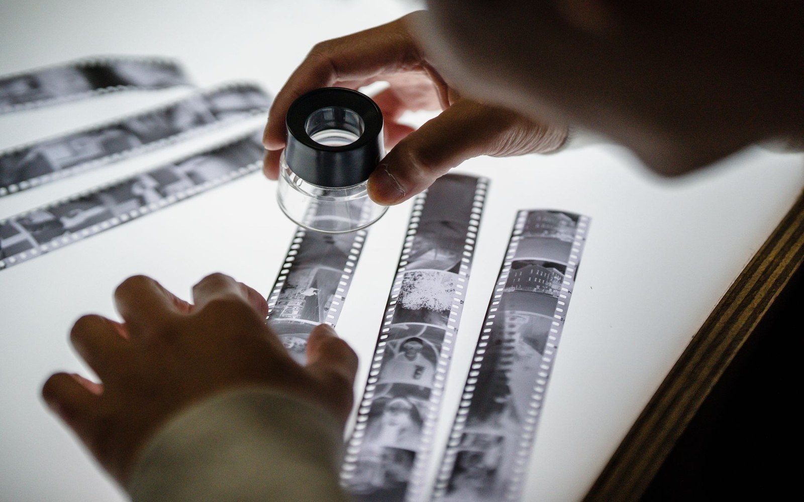 Hands examine black-and-white film negatives with a loupe on a light table