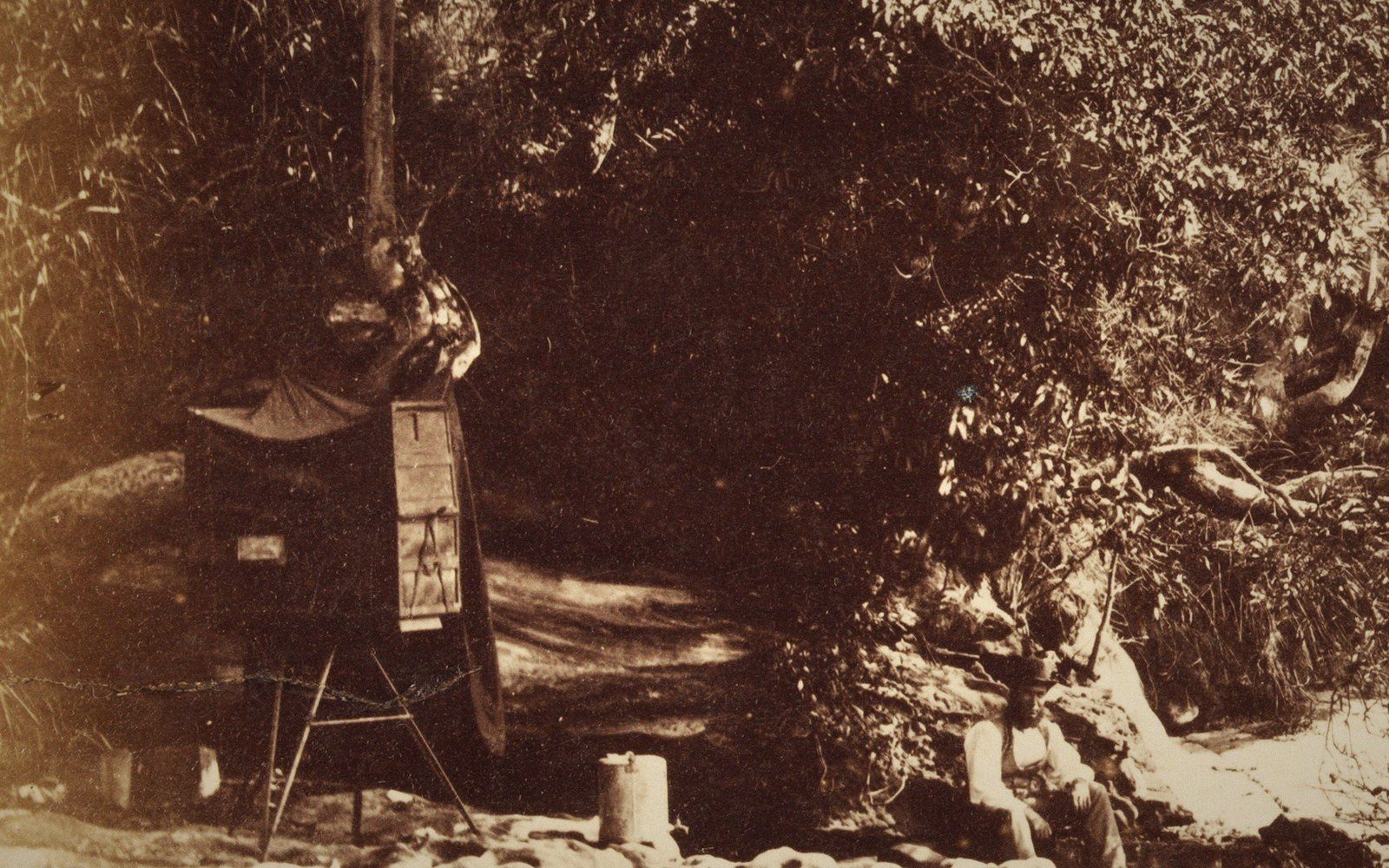 A tented field darkroom on a tripod stands beside a wooded creek with a seated figure