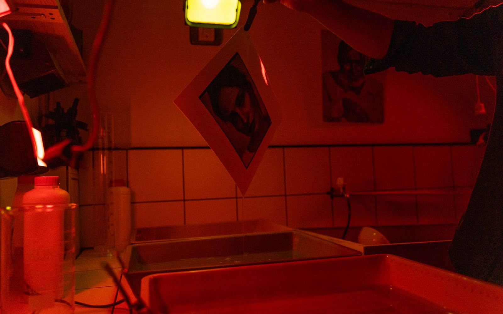 A hand-holding photograph hangs to dry above developing trays in a red-lit darkroom